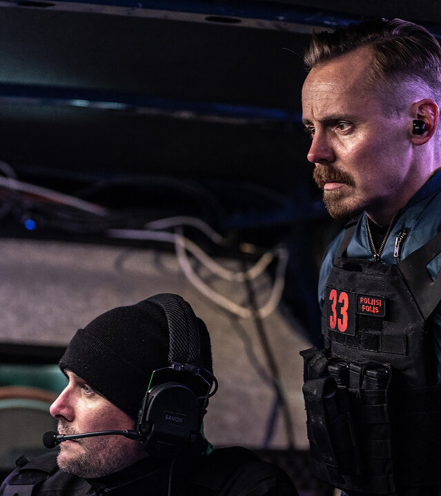 "Operation Omerta - Staatsdiener": Max Tanner (Jasper Pääkkönen) steht in einem Überwachungsfahrzeug neben einem sitzenden Kollegen (Komparse). Beide beobachten etwas. Max trägt eine Schutzweste.