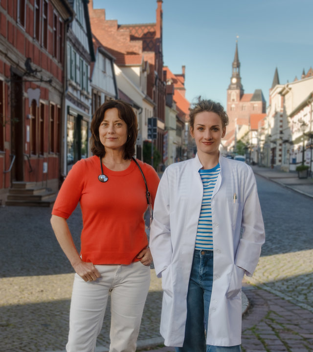 "Mit Herz und Holly - Diagnose Neustart": Neben der Arztpraxis stehen Dr. Katrin Herz (Inka Friedrich) und Dr. Holly Sass (Karoline Teska) auf der Straße. Katrin hat ein Stethoskop um den Hals gelegt, Holly trägt einen weißen Arztkittel. Beide blicken lächelnd in die Kamera.