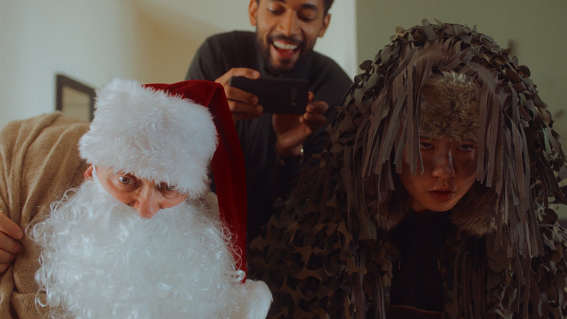 "Doppelhaushälfte - Schöne Bescherung": Andi (Milan Peschel) ist als Weihnachtsmann verkleidet und Rocco (Hoang Minh Ha) als Knecht Ruprecht. Sie schauen nach vorne gebeugt mit intensivem Blick Richtung Kamera. Hinter ihnen steht mich lachendem Blick Theo (Benito Bause) und filmt mit dem Handy das Geschehen.