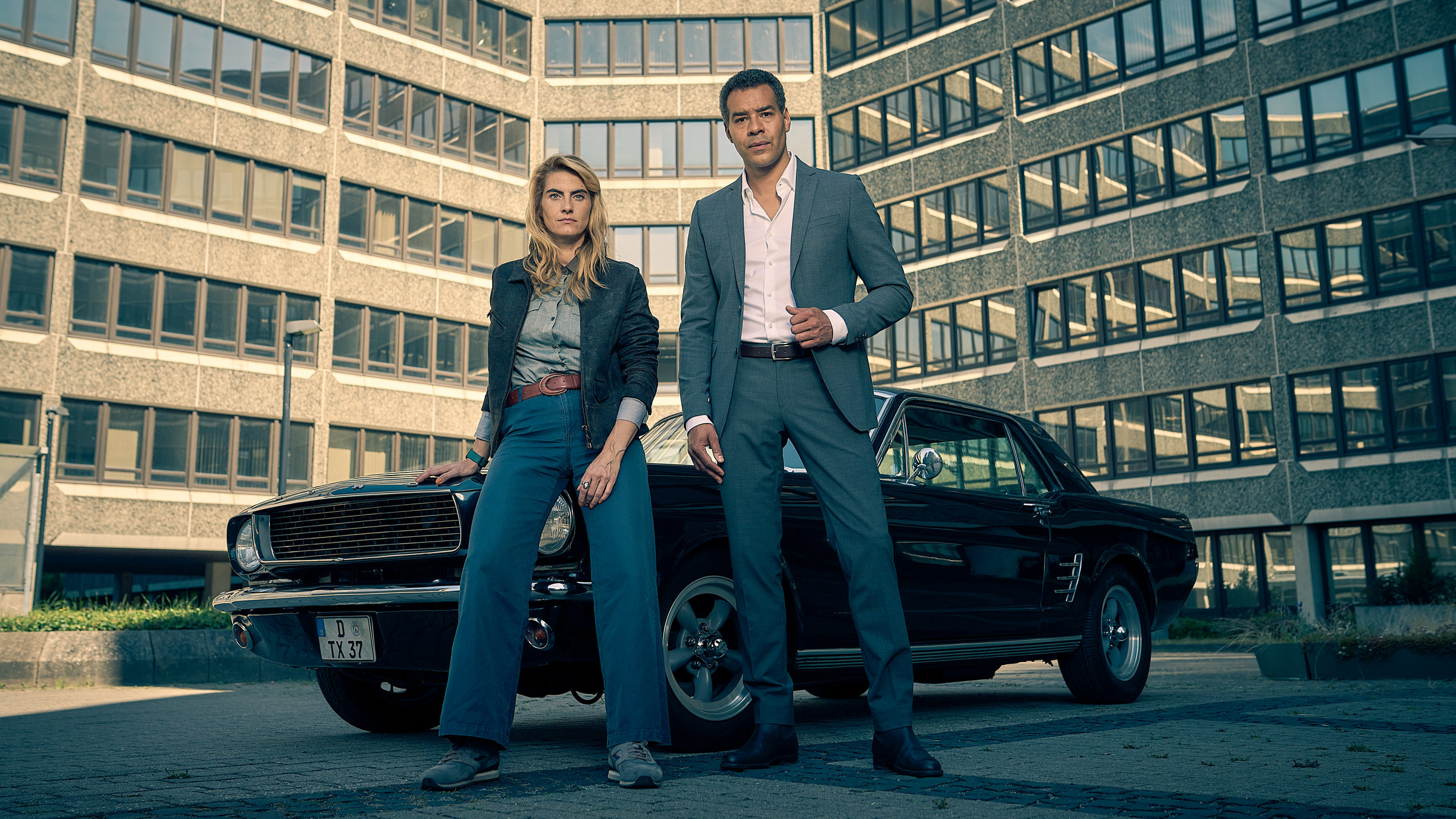 "Unbestechlich": Clarissa Jakobs (Samia Chancrin) lehnt lässig an einem schwarzen Oldtimer. Neben ihr steht Joseph Kanjaa (Michael Klammer) in einem Anzug. Der gesamte Hintergrund wird von einem Bürogebäude ausgefüllt.