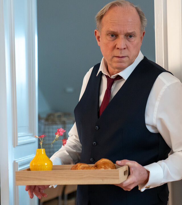 "Letzte Spur Berlin - Fallen Angels": Günther Machmann (Ulrich Tukur) steht, elegant gekleidet, vor einer weißen Holztür. Er hält ein Tablett mit Frühstück in den Händen. Seine Augen sind weit aufgerissen.
