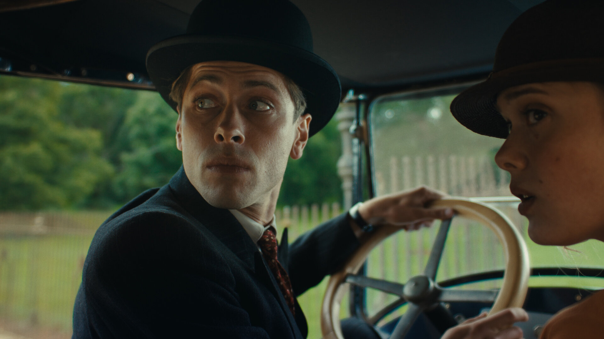 "This Is Not a Murder Mystery - Der Überlebende": René Magritte (Pierre Gervais) sitzt am Steuer eines Autos. Er trägt eine Melone auf dem Kopf. Neben ihm sitzt seine Frau Georgette (Mathilde Warnier), die nur halb im Profil zu sehen ist. Auch sie trägt einen Hut. Beide schauen über die Schulter nach hinten.