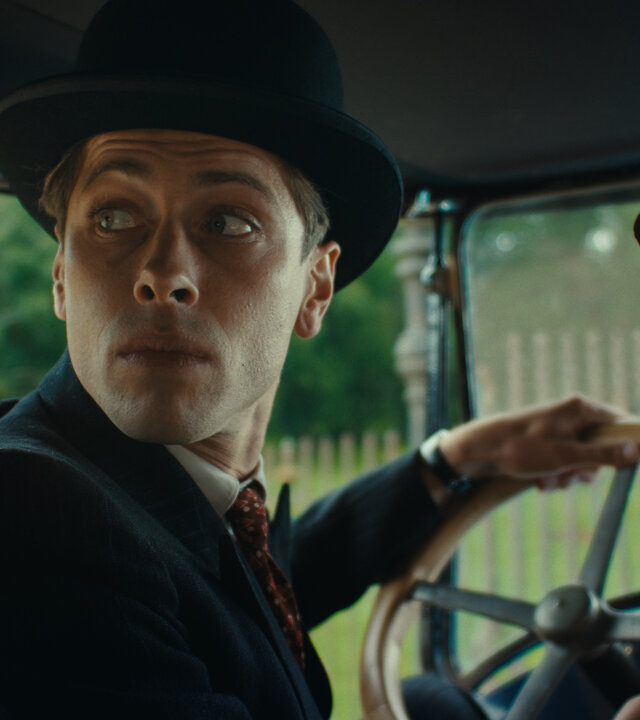 "This Is Not a Murder Mystery - Der Überlebende": René Magritte (Pierre Gervais) sitzt am Steuer eines Autos. Er trägt eine Melone auf dem Kopf. Neben ihm sitzt seine Frau Georgette (Mathilde Warnier), die nur halb im Profil zu sehen ist. Auch sie trägt einen Hut. Beide schauen über die Schulter nach hinten.