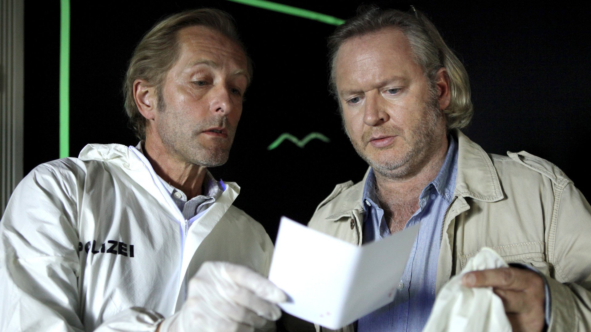 "SOKO Wien - Bis der Vorhang fällt": Spurensicherer Franz Wohlfahrt (Helmut Bohatsch) informiert Oberstleutnant Helmuth Nowak (Gregor Seberg) über die neuesten Spurenerkenntnisse. Franz Wohlfahrt trägt den weißen Spurensicherungsoverall mit der Aufschrift "Polizei" und Gummihandschuhe. In der rechten Hand hält er einen Zettel, den er Nowak zeigt. Nowak trägt ein blaues Hemd mit heller Jeansjacke. In der linken Hand hält er Einweghandschuhe und schaut auf das Blatt Papier.