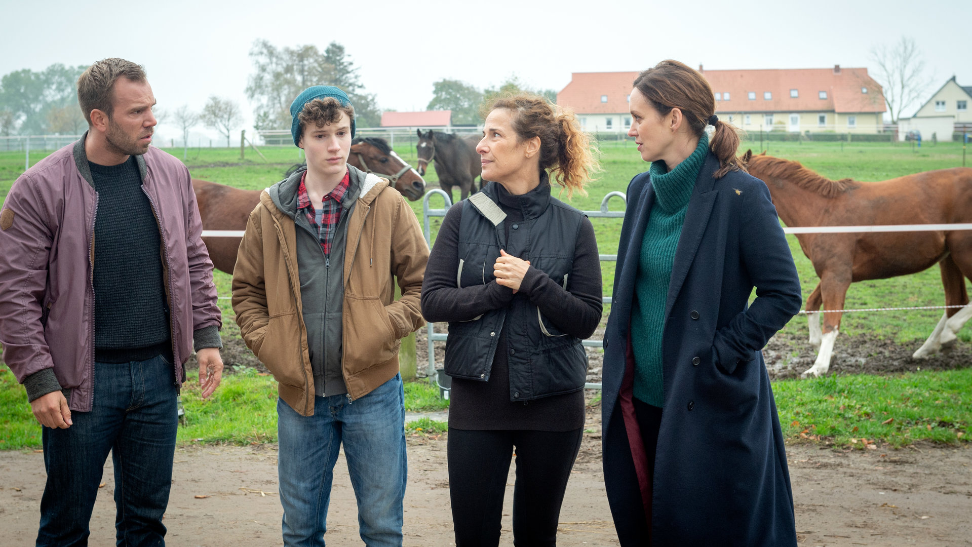 "SOKO Wismar – Das Leben ist kein Ponyhof": Die Kommissare Pöhlmann (Dominic Boeer) und Joost (Nike Fuhrmann) stehen mit Gesine (Clelia Sarto) und Jonathan von Gungelitz (Nico Ramon Kleemann) vor einer begrünten Pferdekoppel. Einige Pferde galoppieren auf der Koppel. Etwas entfernt im Hintergrund liegt ein Gästehaus.
