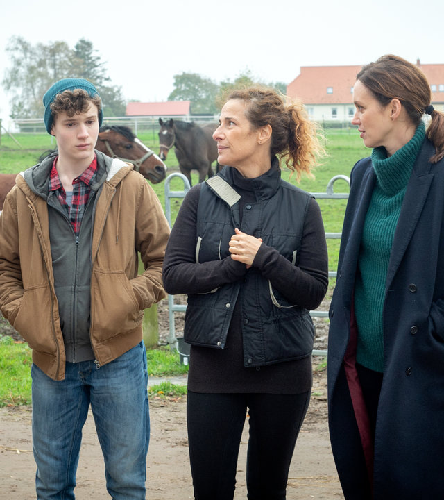 "SOKO Wismar – Das Leben ist kein Ponyhof": Die Kommissare Pöhlmann (Dominic Boeer) und Joost (Nike Fuhrmann) stehen mit Gesine (Clelia Sarto) und Jonathan von Gungelitz (Nico Ramon Kleemann) vor einer begrünten Pferdekoppel. Einige Pferde galoppieren auf der Koppel. Etwas entfernt im Hintergrund liegt ein Gästehaus.