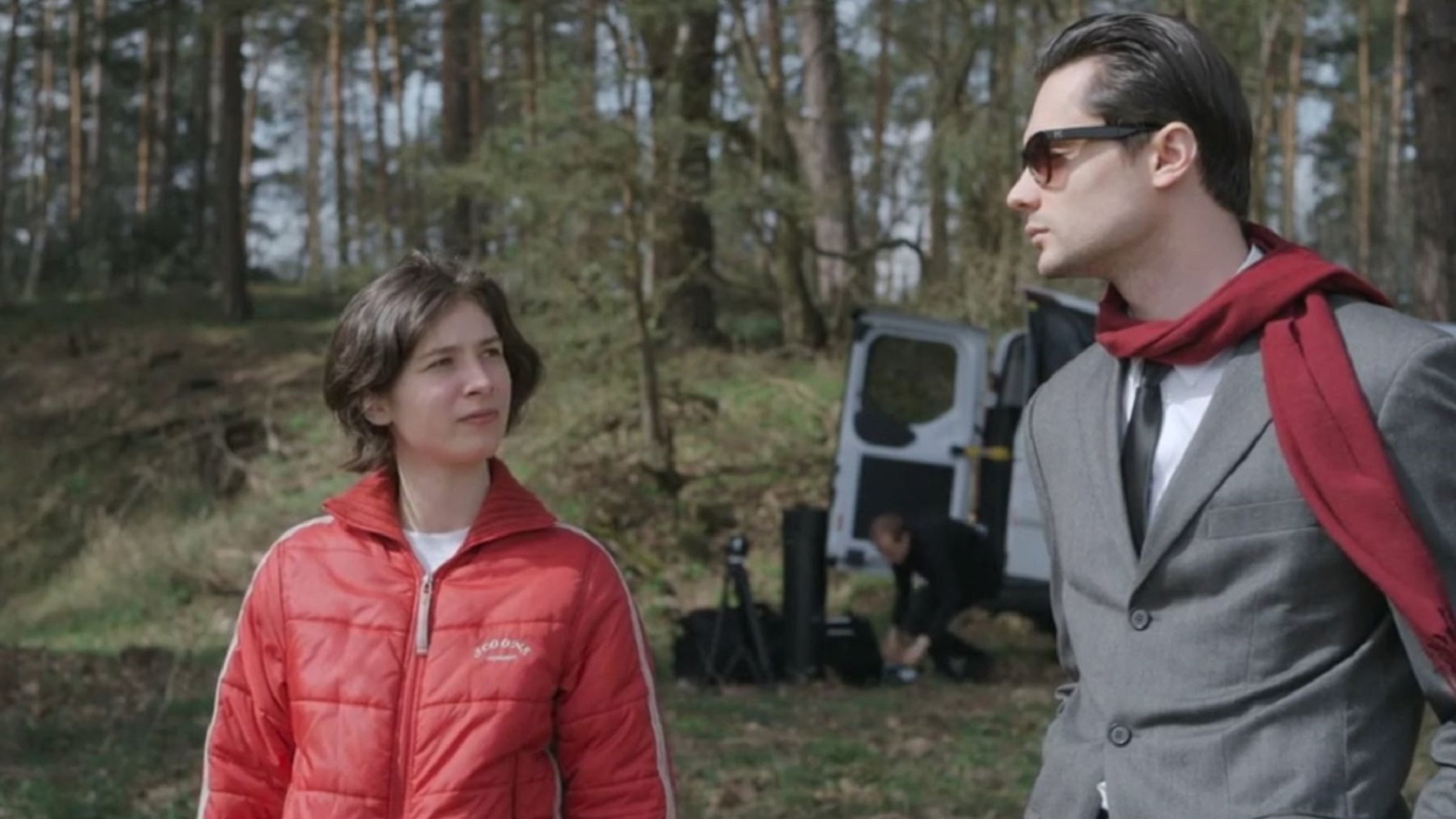 "Which Way to the West": An einem Waldrand, im Hintergrund ein geöffneter Minitrransporter, aus dem ein Mann Filmausrüstung auspackt: Eine junge Frau mit halblangen dunkelblonden Haaren in einem roten Anorak schaut prüfend einen schick angezogenen jungen Mann an, der in grauem Jackett mit weißem Hemd und schwarzer Krawatte, einen roten Wollschal um den Hals, durch seine schwarze Sonnenbrille distanziert auf sie herabblickt.