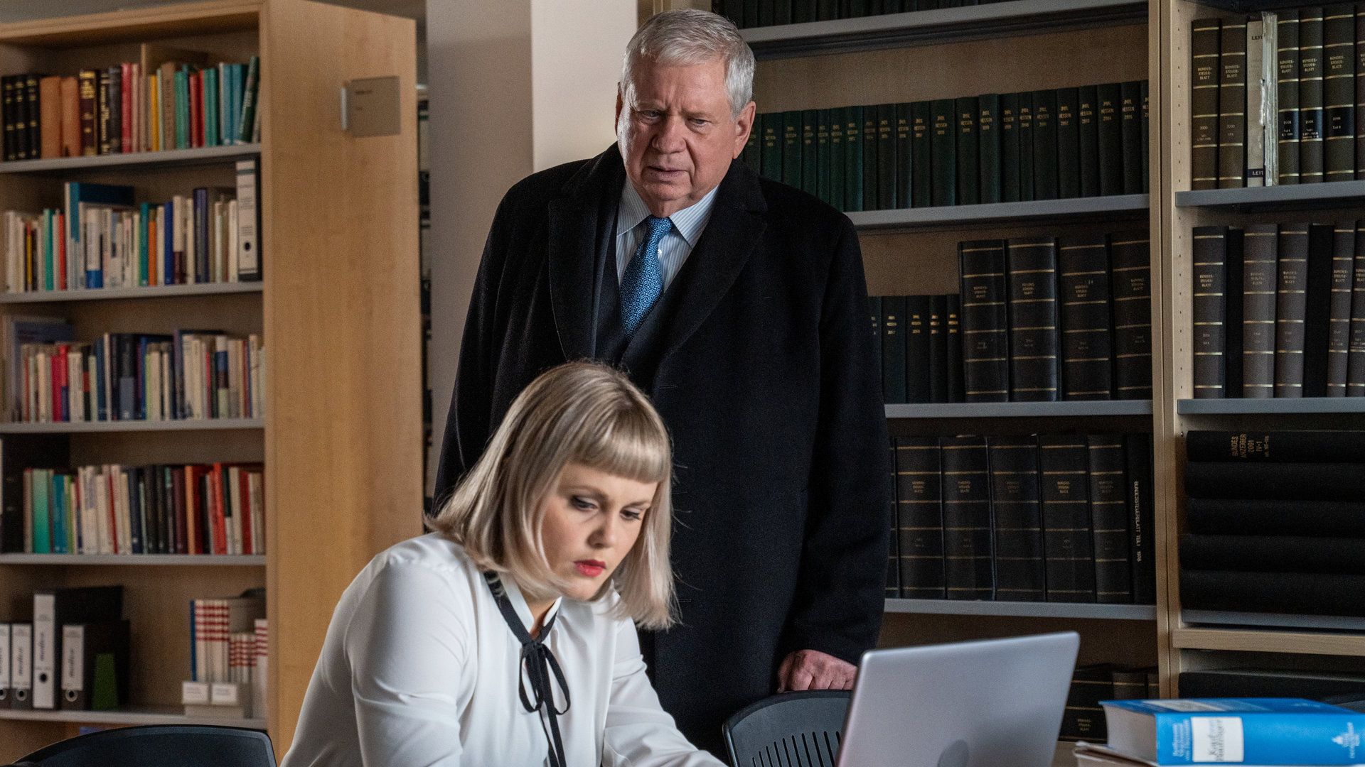 "Der Staatsanwalt - Unter Druck": Bernd Reuther (Rainer Hunold) steht in einer Bibliothek neben einem Schreibtisch, an dem Dr. Alina Berg (Paulina Alpen) sitzt. Berg arbeitet an einem Laptop. Vor ihr auf dem Schreibtisch liegen verschiedene Bücher. Reuther blickt skeptisch auf den Bildschirm des Laptops.