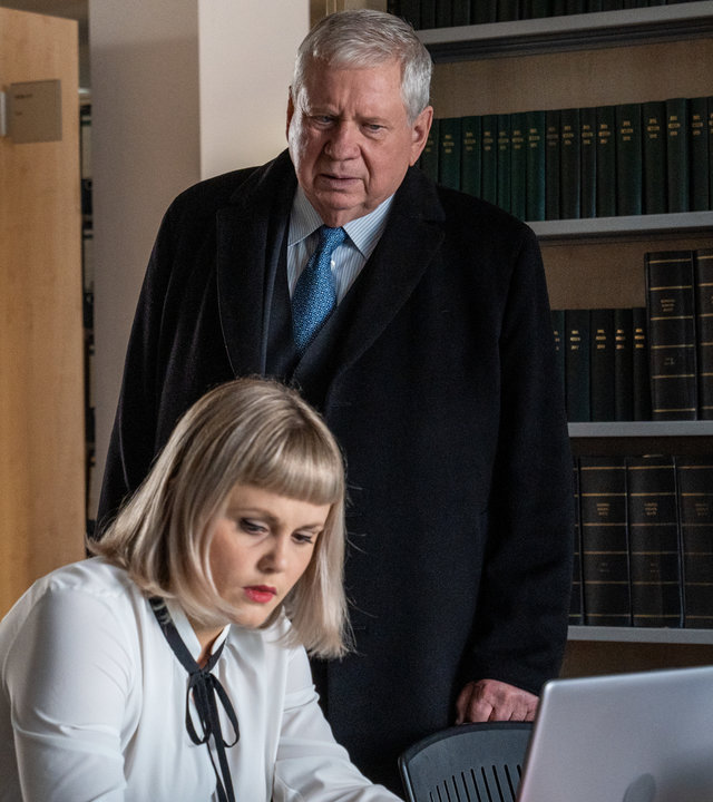 "Der Staatsanwalt - Unter Druck": Bernd Reuther (Rainer Hunold) steht in einer Bibliothek neben einem Schreibtisch, an dem Dr. Alina Berg (Paulina Alpen) sitzt. Berg arbeitet an einem Laptop. Vor ihr auf dem Schreibtisch liegen verschiedene Bücher. Reuther blickt skeptisch auf den Bildschirm des Laptops.