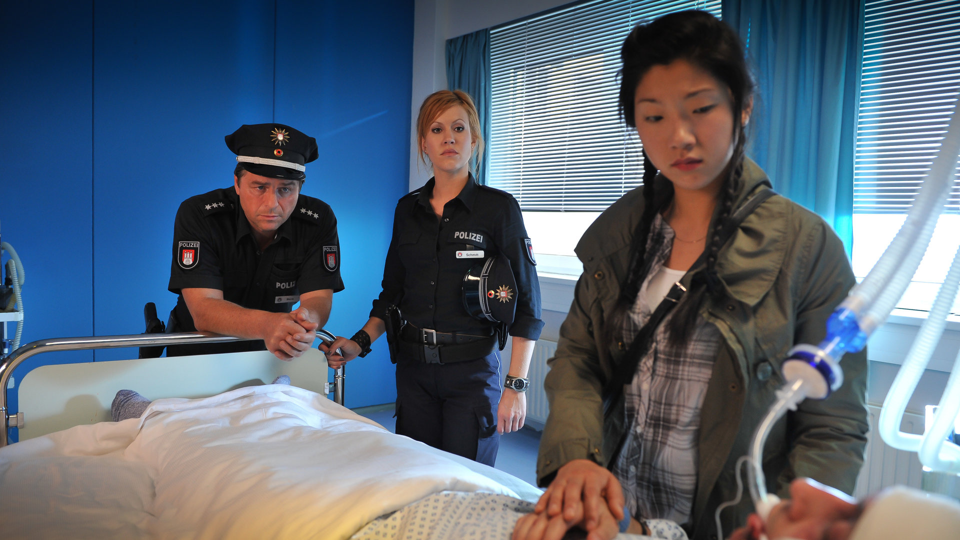 "Notruf Hafenkante - Heiße Ware": Maggie Li (Joy Maria Bai) steht an einem Bett im Krankenhaus und hält die Hand des darin liegenden Patienten. Am Fußende des Bettes stehen Jule (Wolke Hegenbarth) und Henning (Uwe Fellensiek) und beobachten sie skeptisch.
