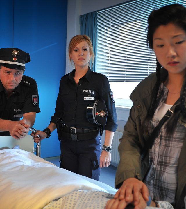 "Notruf Hafenkante - Heiße Ware": Maggie Li (Joy Maria Bai) steht an einem Bett im Krankenhaus und hält die Hand des darin liegenden Patienten. Am Fußende des Bettes stehen Jule (Wolke Hegenbarth) und Henning (Uwe Fellensiek) und beobachten sie skeptisch.