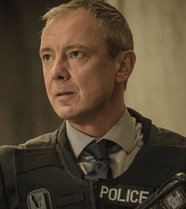 "Detective Grace - Du sollst nicht sterben": DS Roy Grace (John Simm) in Nahaufnahme: Er schaut entschlossen zur Seite und trägt eine schusssichere Weste mit der Aufschrift "POLICE" über seinem Anzug.