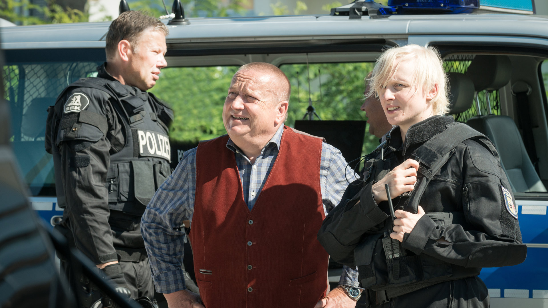 "SOKO Wismar – Überfall!": Revierchef Reuter (Udo Kroschwald) steht zwischen einem SEK-Kollegen (Komparse) und der uniformierten SEK-Beamtin Elke Brandt (Anne Müller) vor einem blauen Polizeibus.