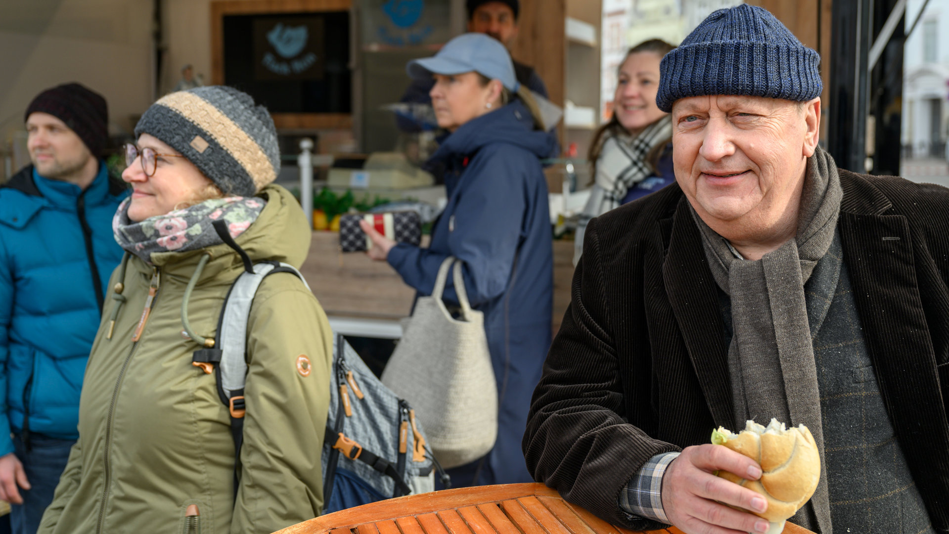 "SOKO Wismar – Marktschreier": Vor einer Bude auf dem Marktplatz steht Jan-Hinrich Reuter (Udo Kroschwald) und isst ein Fischbrötchen. Um ihn herum stehen Besucher des Marktes. Reuter hört amüsiert den Marktschreiern zu.