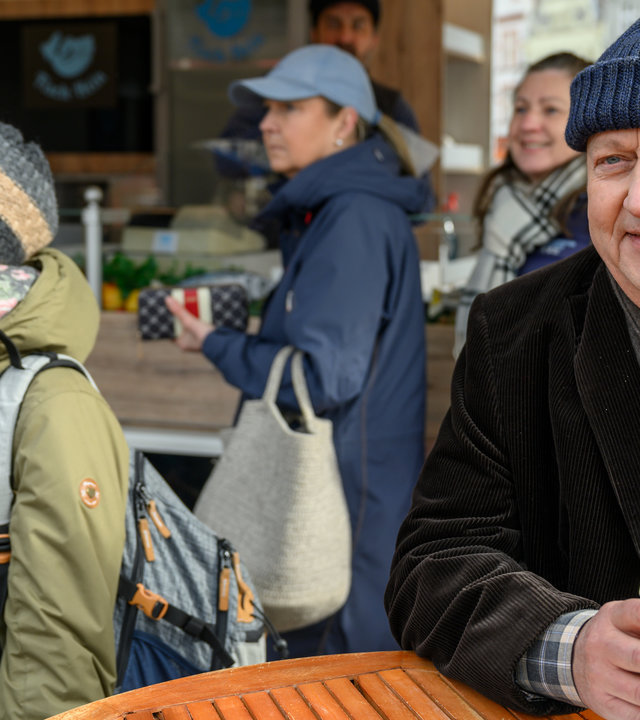 "SOKO Wismar – Marktschreier": Vor einer Bude auf dem Marktplatz steht Jan-Hinrich Reuter (Udo Kroschwald) und isst ein Fischbrötchen. Um ihn herum stehen Besucher des Marktes. Reuter hört amüsiert den Marktschreiern zu.