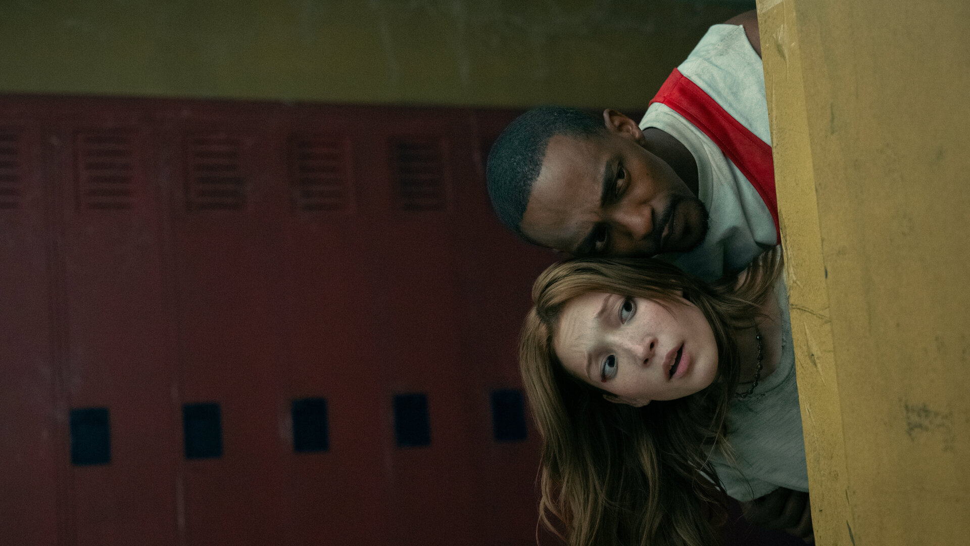"Twisted Metal - SDDNDTH": Mayhem (Saylor Bell Curda) und John (Anthony Mackie) verstecken sich hintereinander. In gebückter Haltung und mit eng zusammengesteckten Köpfen blicken sie nach vorne. Während Mayhem verängstigt nach vorne schaut, blickt John konzentriert auf das Geschehen. Im Hintergrund befindet sich eine Reihe roter Schulspinde.