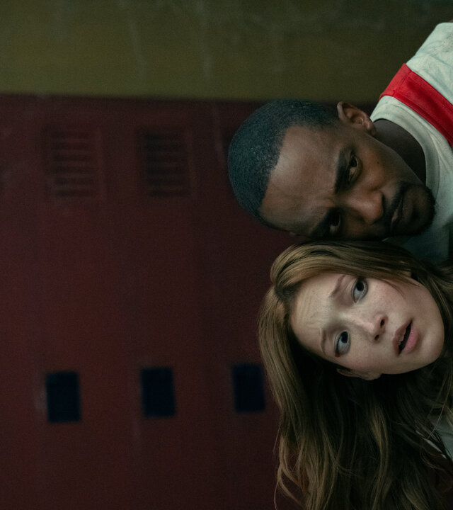 "Twisted Metal - SDDNDTH": Mayhem (Saylor Bell Curda) und John (Anthony Mackie) verstecken sich hintereinander. In gebückter Haltung und mit eng zusammengesteckten Köpfen blicken sie nach vorne. Während Mayhem verängstigt nach vorne schaut, blickt John konzentriert auf das Geschehen. Im Hintergrund befindet sich eine Reihe roter Schulspinde.