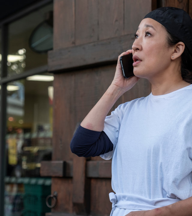 "Killing Eve - Im Exil": Eve (Sandra Oh) steht vor einem Gebäude und telefoniert. Dabei trägt sie ein weißes T-Shirt und eine Schürze.