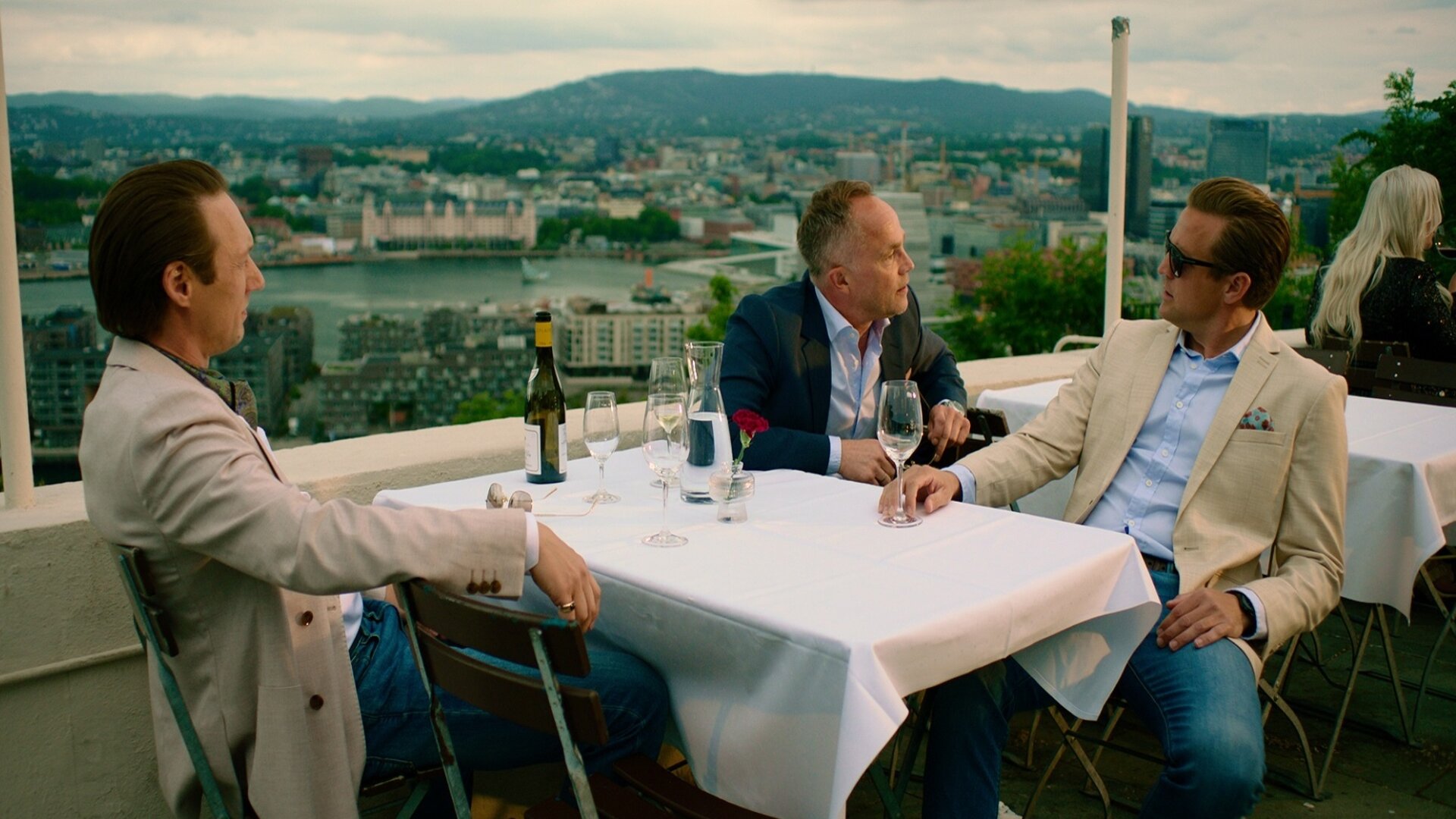 "Exit - Family first": Adam (Simon J. Berger), Jeppe (Jon Øigarden) und Henrik (Tobias Santelmann) sitzen niedergeschlagen an einem Restauranttisch im Freien. Auf dem Tisch stehen eine Flasche Wein und Weingläser.
