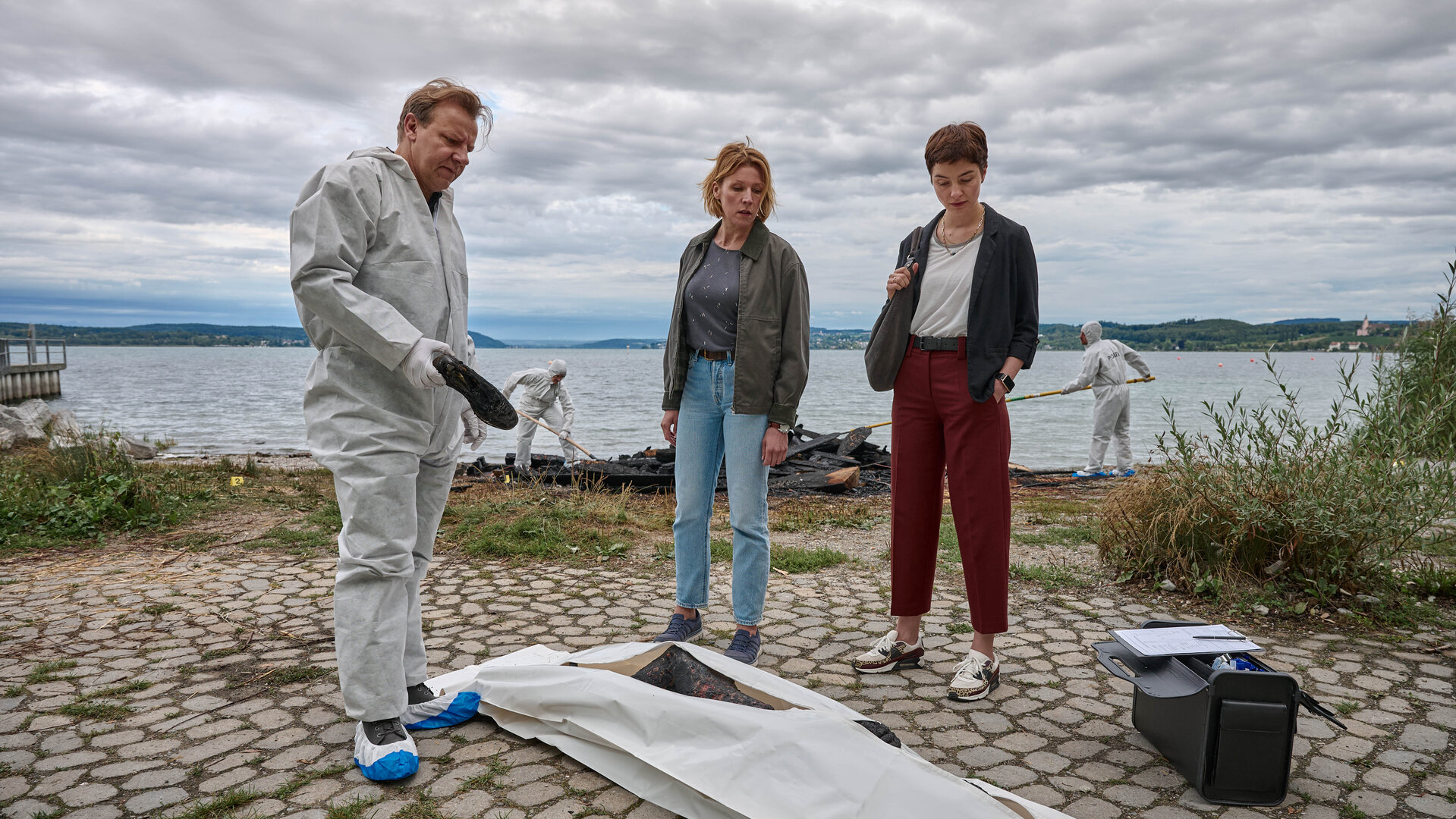 "Mordufer - Brennen soll sie": Gregor Eisen (Ronald Kukulies), Doro Beitinger (Franziska Weisz) und Chiara Locatelli (Maria Wördemann) stehen am Ufer des Bodensees an einem Tatort. Vor ihnen auf dem Boden liegt eine Brandleiche in einem Leichensack. Im Hintergrund sind Spurensicherer bei der Arbeit und suchen das Ufer ab. Gregor Eisen und die Spurensicherer tragen weiße Overalls, blau-weiße Überzieher über ihren Schuhen und weiße Handschuhe.