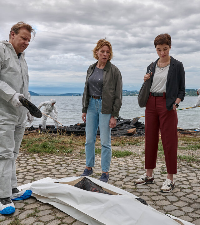 "Mordufer - Brennen soll sie": Gregor Eisen (Ronald Kukulies), Doro Beitinger (Franziska Weisz) und Chiara Locatelli (Maria Wördemann) stehen am Ufer des Bodensees an einem Tatort. Vor ihnen auf dem Boden liegt eine Brandleiche in einem Leichensack. Im Hintergrund sind Spurensicherer bei der Arbeit und suchen das Ufer ab. Gregor Eisen und die Spurensicherer tragen weiße Overalls, blau-weiße Überzieher über ihren Schuhen und weiße Handschuhe.