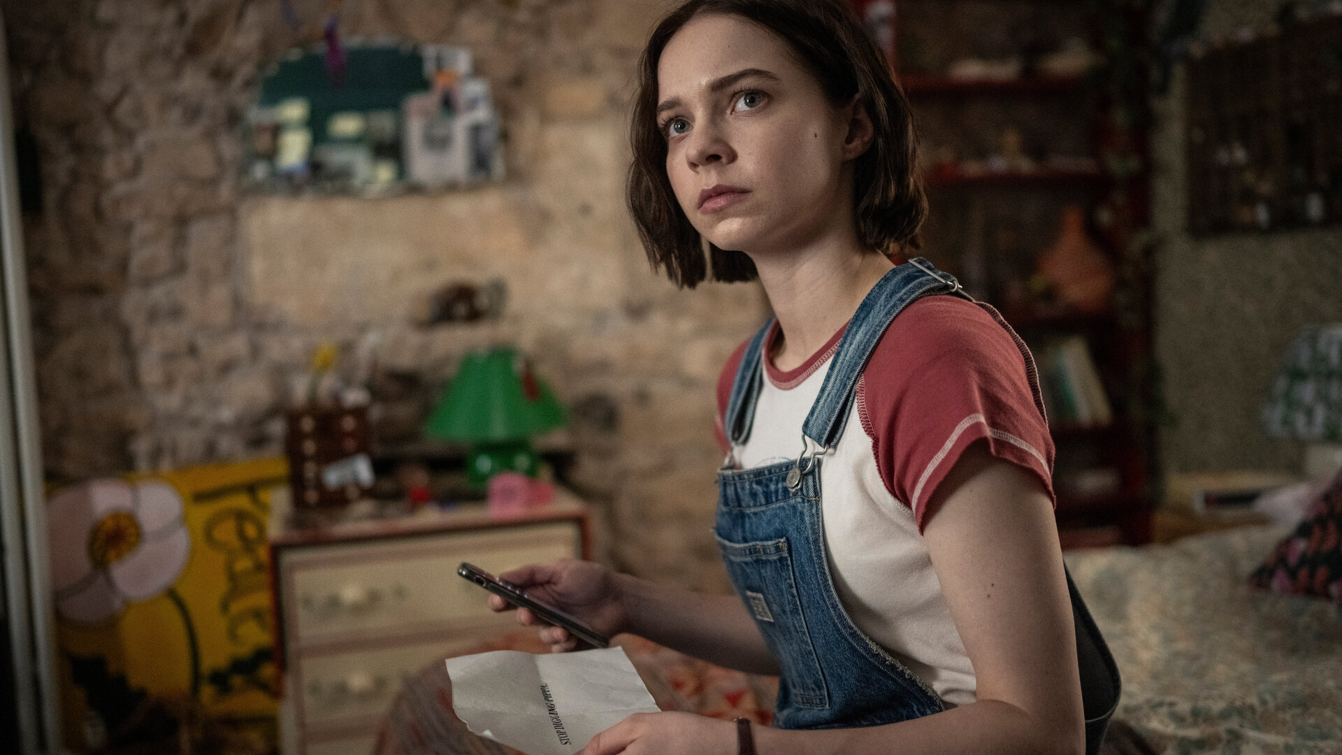 "A Good Girl's Guide to Murder – Hör auf zu graben, Pippa.": Pip (Emma Myers) sitzt auf ihrem Bett, sie trägt eine blaue Latzhose über einem rot weißen T-Shirt. In der rechten Hand hält sie ein Handy, in der linken Hand einen Zettel mit einer Drohbotschaft. Sie schaut besorgt.