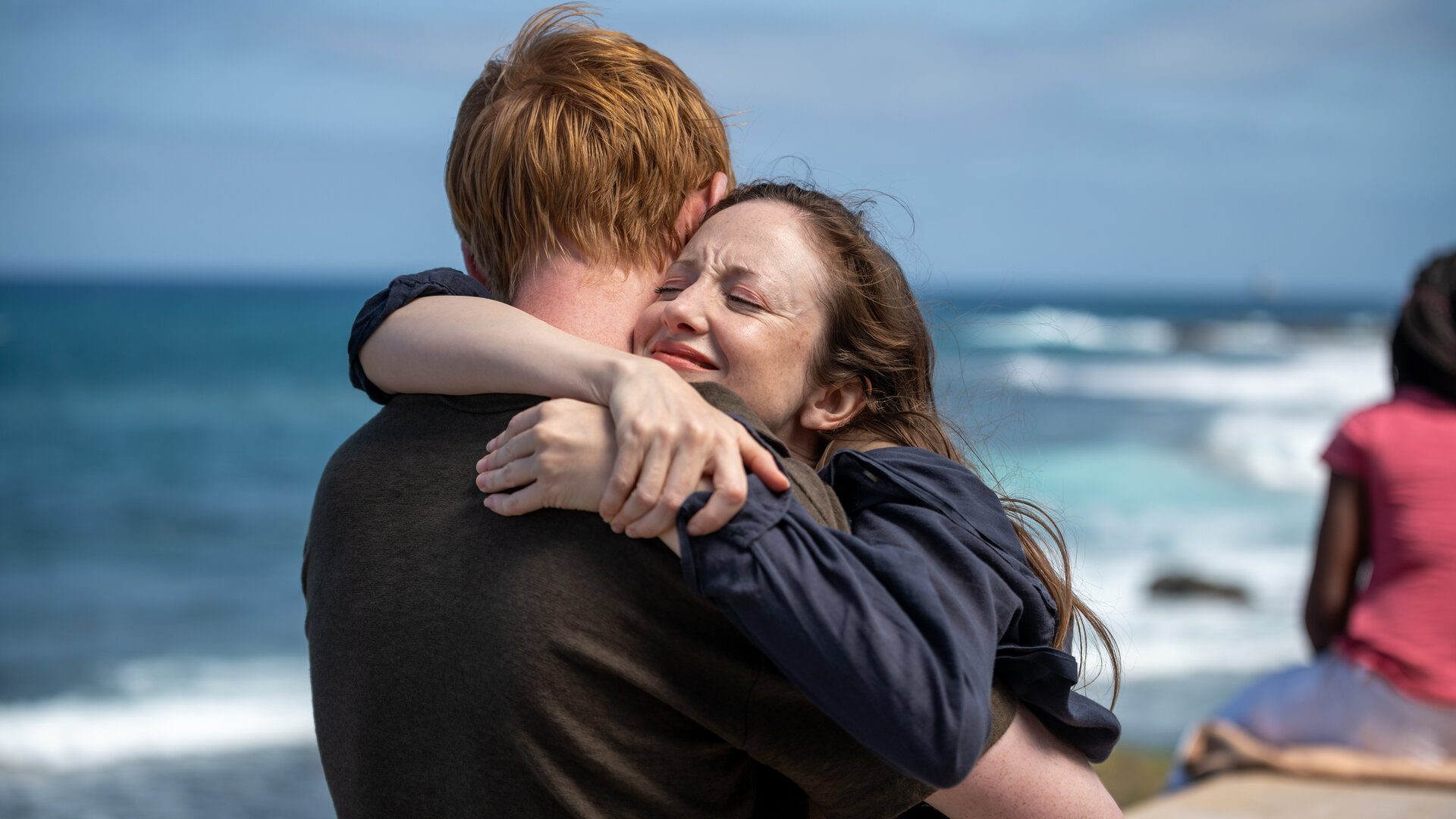 "Alice & Jack - Wiedersehen": Jack (Domhnall Gleeson) hält Alice (Andrea Riseborough) fest im Arm. Sie stehen am Meer, hinter ihnen schlägt das Meer Wellen und der Himmel ist blau.