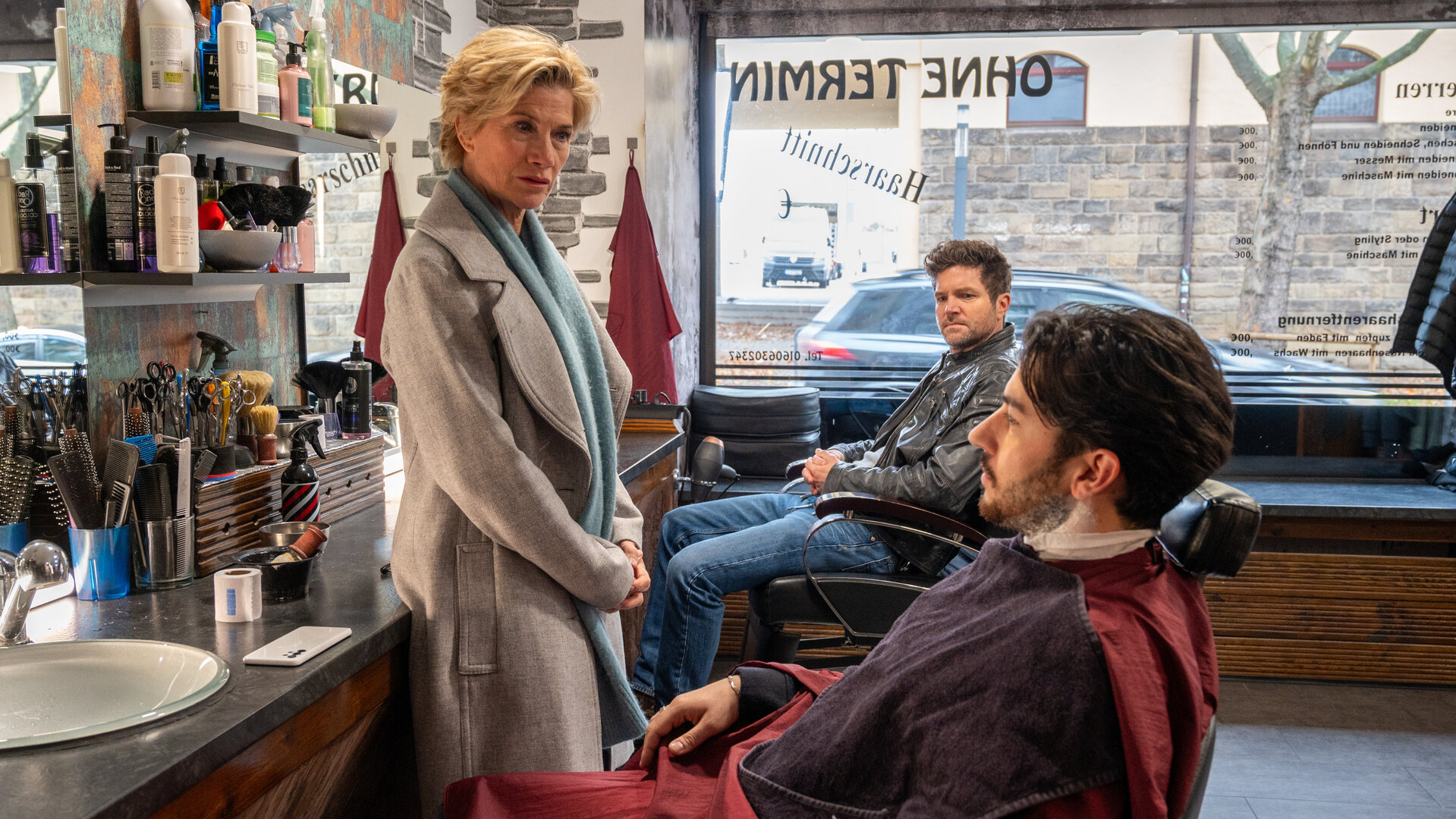 "SOKO Stuttgart - Sündenfall im Vatikan": In einem Friseurladen sitzt Gino de Luca (David Brizzi) mit einem Friseurumhang auf einem Friseurstuhl. Neben ihm steht die Kommissarin Martina Seiffert (Astrid M. Fünderich) und schaut ihn ernst an. Im Hintergrund sitzt Kommissar Jo Stoll (Peter Ketnath) auf einem weiteren Friseurstuhl und schaut kritisch auf Gino.