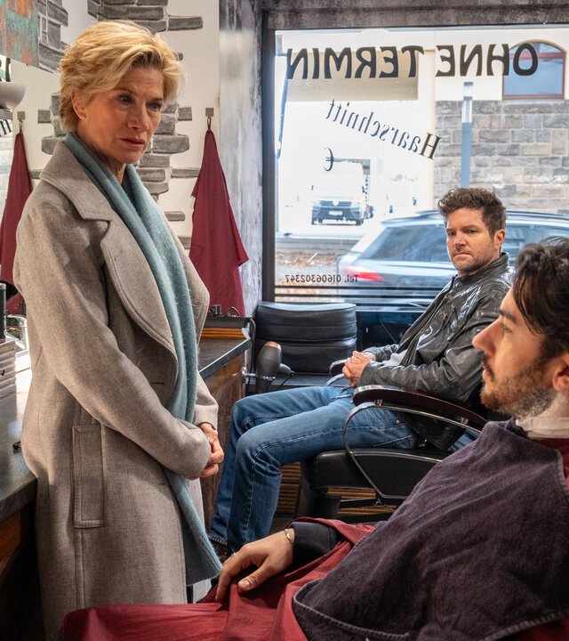 "SOKO Stuttgart - Sündenfall im Vatikan": In einem Friseurladen sitzt Gino de Luca (David Brizzi) mit einem Friseurumhang auf einem Friseurstuhl. Neben ihm steht die Kommissarin Martina Seiffert (Astrid M. Fünderich) und schaut ihn ernst an. Im Hintergrund sitzt Kommissar Jo Stoll (Peter Ketnath) auf einem weiteren Friseurstuhl und schaut kritisch auf Gino.