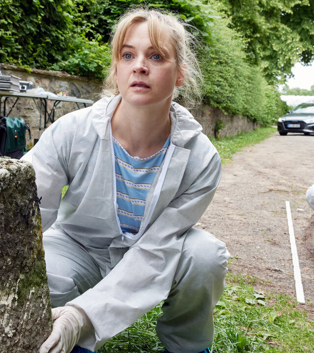 "Mordsschwestern - Verbrechen ist Familiensache: Das Chamäleon": Feli (Caroline Hanke) und Dusi (Claudiu Mark Draghici) sind am Tatort. Feli hockt vor einem moosbewachsenen Stein auf dem sich sich ein gelbes Schild mit der Zahl fünf befindet. Dusi hockt etwas weiter hinter Feli und hält eine Kamera in den Händen. Beide tragen weiße Schutzanzüge und Gummihandschuhe.