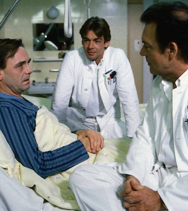 "Die Schwarzwaldklinik - Die Wunderquelle": Dr. Rens (Holger Petzold) liegt im Krankenbett. Professor Brinkmann (Klausjürgen Wussow) und sein Sohn Udo (Sascha Hehn) stehen neben seinem Bett und reden mit ihm.