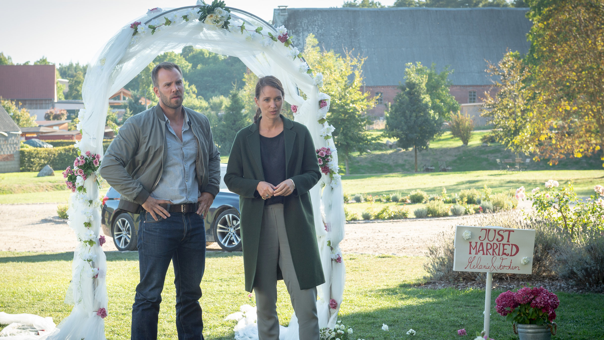 "SOKO Wismar - Verliebt, verlobt, verstorben": Lars Pöhlmann (Dominic Boeer) und Karoline Joost (Nike Fuhrmann) stehen im Grünen vor einem, mit Rosen verzierten, Hochzeitsbogen. Lars hat entschieden die Arme in die Hüften gestemmt, Karoline hält ihre Arme angewinkelt vorm Körper. Ein Blumenkübel mit Hortensien steht herum. Daneben ist ein Schild mit der Aufschrift „Just Married“ in den Boden gesteckt. Im Hintergrund liegt das stattliche Anwesen der Bremers.