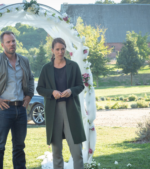 "SOKO Wismar - Verliebt, verlobt, verstorben": Lars Pöhlmann (Dominic Boeer) und Karoline Joost (Nike Fuhrmann) stehen im Grünen vor einem, mit Rosen verzierten, Hochzeitsbogen. Lars hat entschieden die Arme in die Hüften gestemmt, Karoline hält ihre Arme angewinkelt vorm Körper. Ein Blumenkübel mit Hortensien steht herum. Daneben ist ein Schild mit der Aufschrift „Just Married“ in den Boden gesteckt. Im Hintergrund liegt das stattliche Anwesen der Bremers.