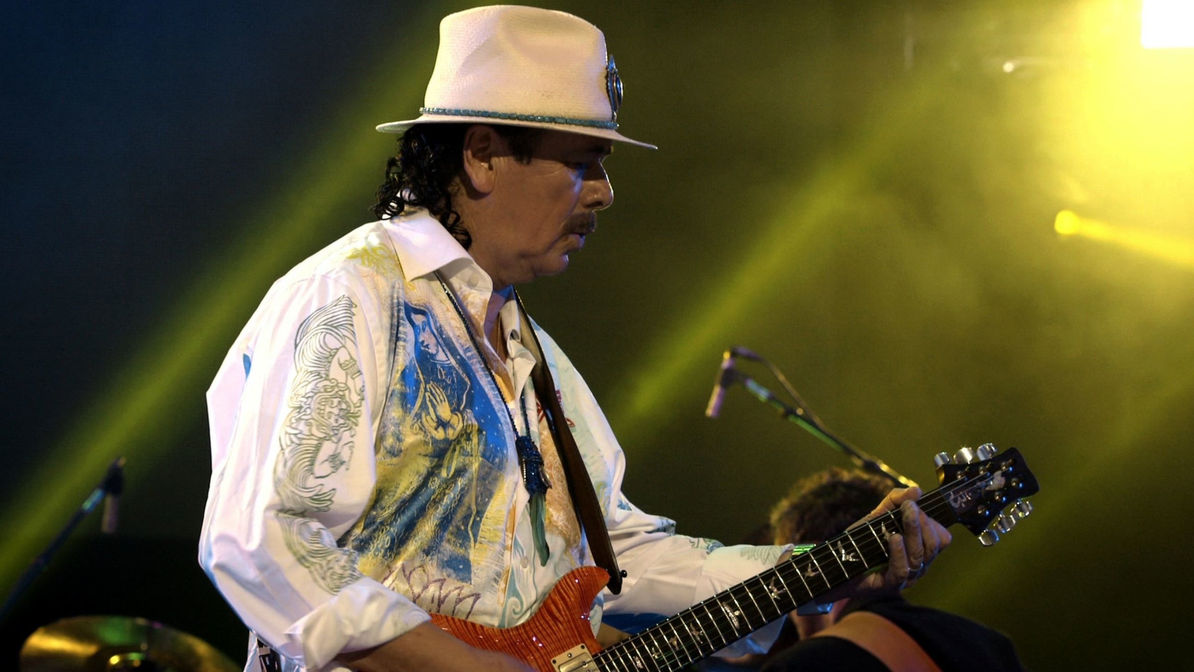 "Santana: Corazón - Live from Mexico": Carlos Santana während eines Konzerts auf der Bühne. Er spielt Gitarre. Bekleidet ist er mit einem weißen Hemd mit auffälligem Muster. Er trägt einen weißen Hut.