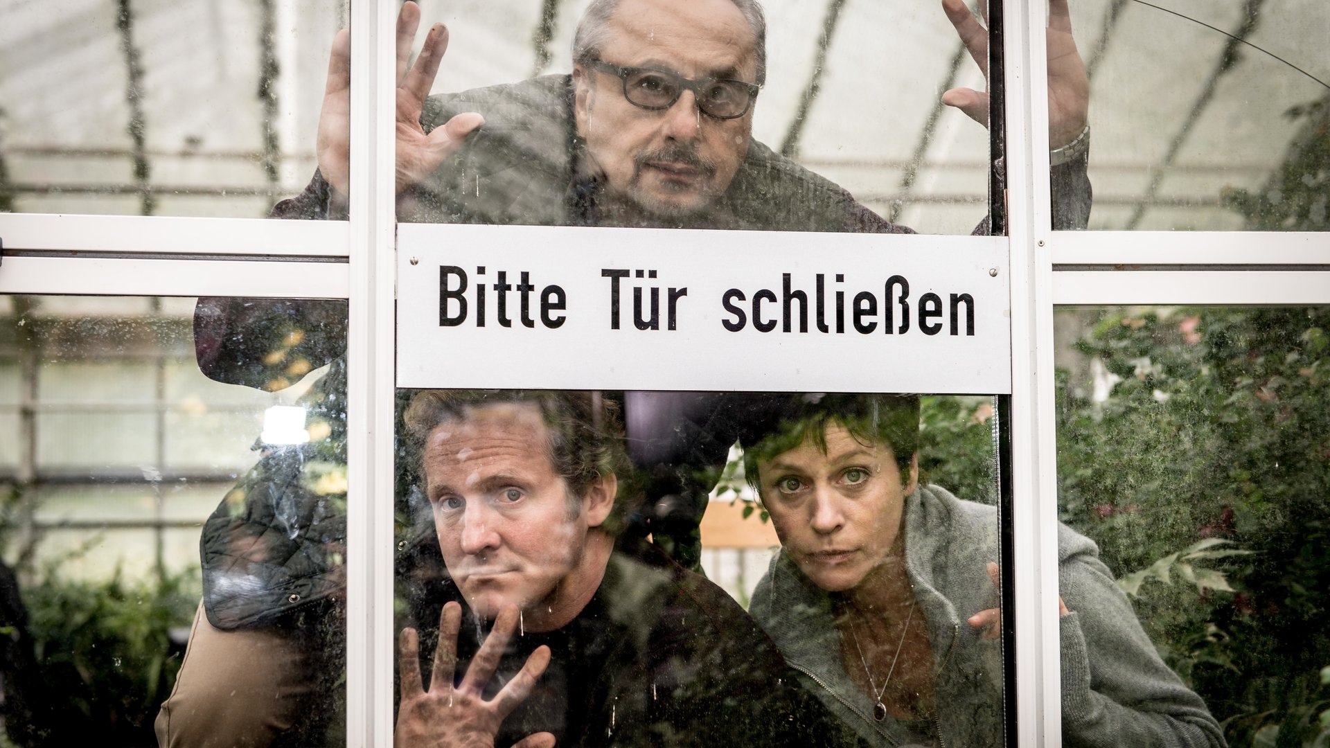 "Die Insassen": Löhring (Wolfgang Stumph), Winter (Maximilian Brückner) und Karin Schlick (Jule Ronstedt, hinter der Tür eines Glasgewächshauses, auf der steht "Bitte Tür schließen".
