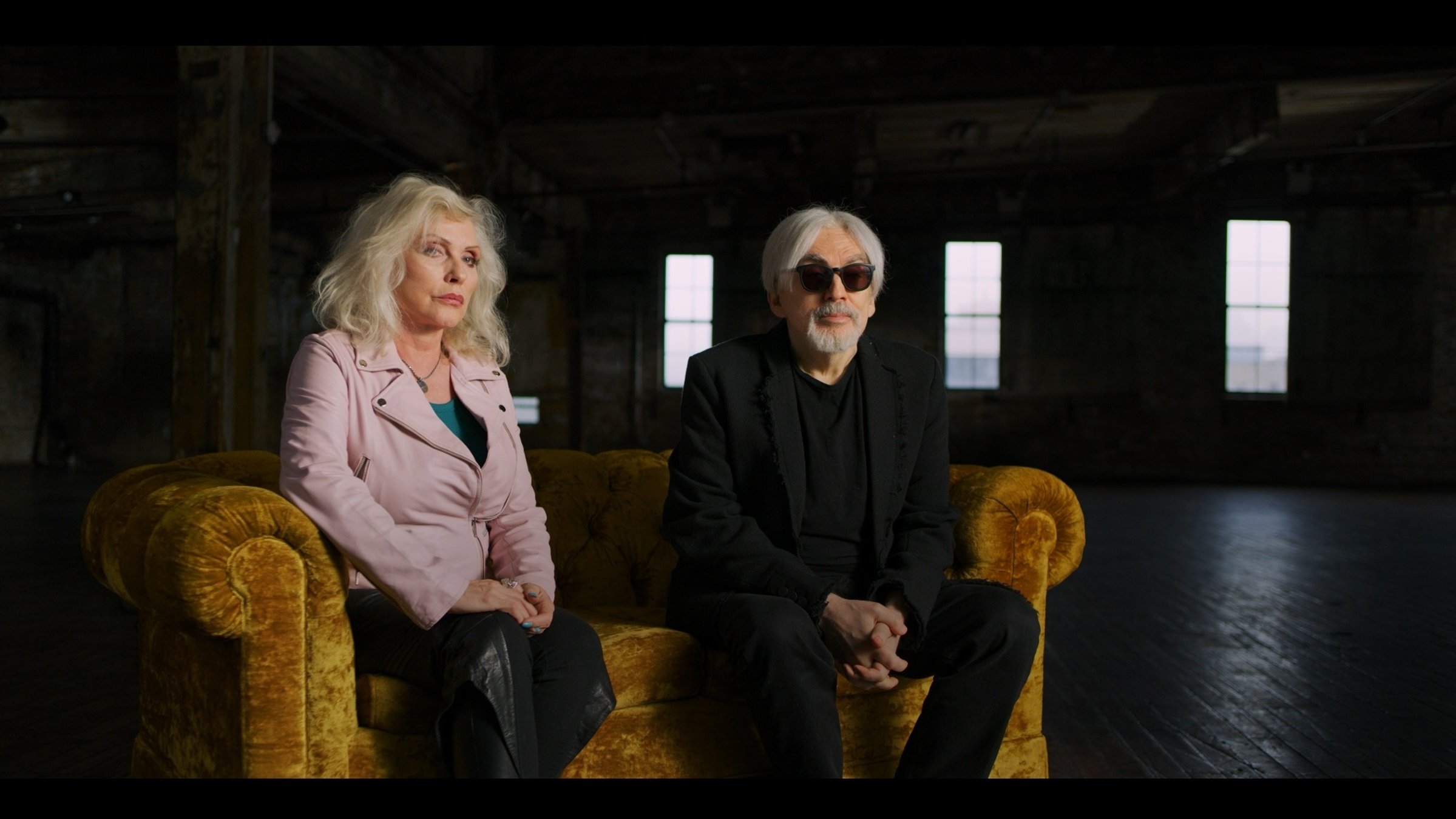 "The True Story of Punk - Die Geburt": Sängerin Debbie Harry und Gitarrist Chris Stein sitzen auf einem ockerfarbenen Sofa in einer Interviewsituation.