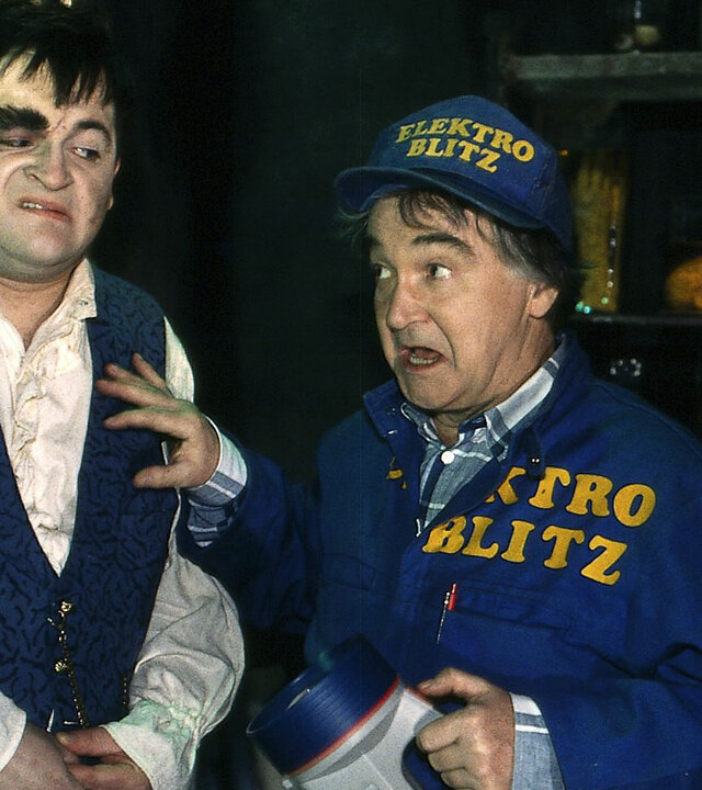 "Sprechstunde bei Dr. Frankenstein - Vorsicht, Killertomaten!": Lenni (Michael Trischan) steht mit dem Elektriker (Victor Deiß) in einem dunklen Raum. Lenni schaut skeptisch zum Elektriker, dieser blickt mit weit aufgerissenen Augen seitlich nach vorne.