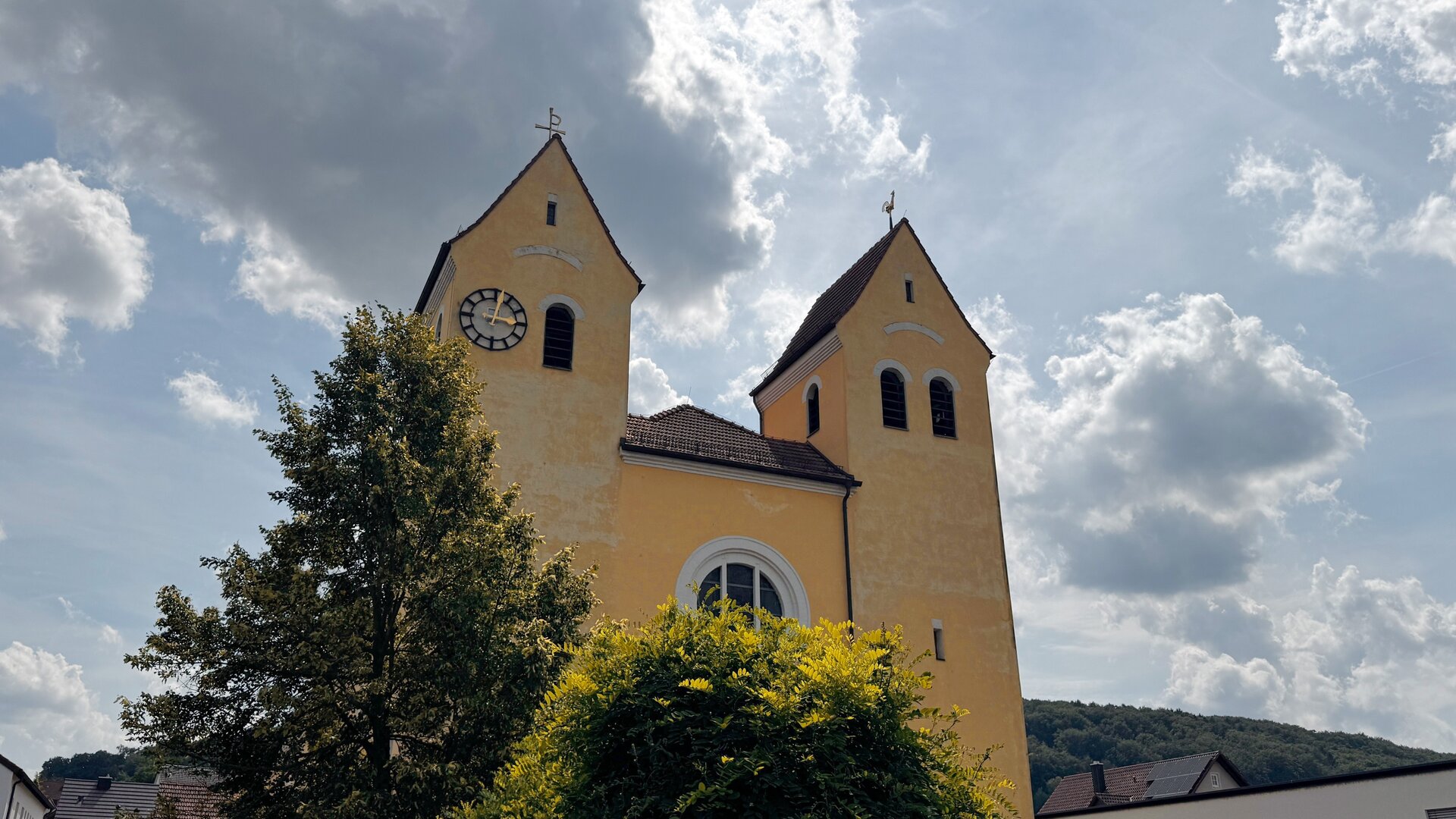 "Katholischer Gottesdienst - Mach's einfach!": Untersicht, Außenaufnahme der Kirche Heilig Kreuz in Kelheim: Zwei Türme der Kirche ragen in den wolkenverhangenen Himmel, die Sonne durchbricht teilweise die Wolkendecke.