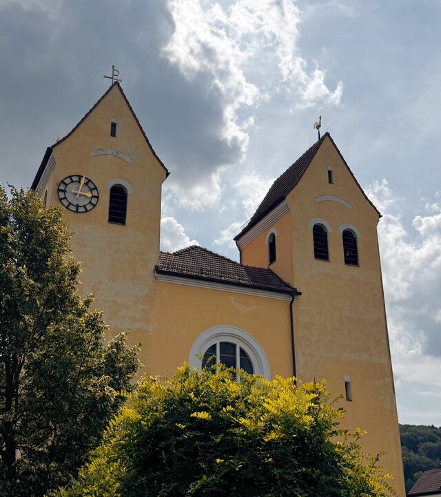 "Katholischer Gottesdienst - Mach's einfach!": Untersicht, Außenaufnahme der Kirche Heilig Kreuz in Kelheim: Zwei Türme der Kirche ragen in den wolkenverhangenen Himmel, die Sonne durchbricht teilweise die Wolkendecke.