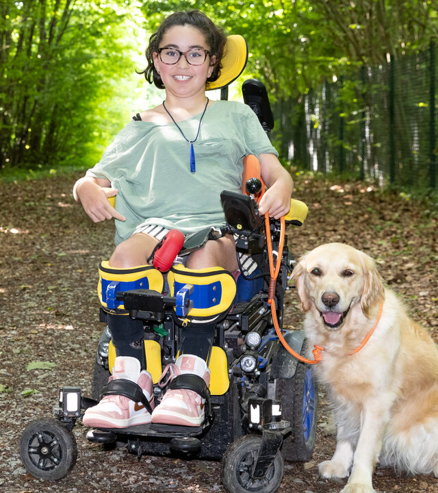 "einfach Mensch - Lena Staudt: Hund Ypsi in meiner Welt": Die 12-jährige Lena sitzt im Wald im Rollstuhl, ihre Assistenzhündin Ypsi sitzt daneben. Lena schaut lächelnd in die Kamera.