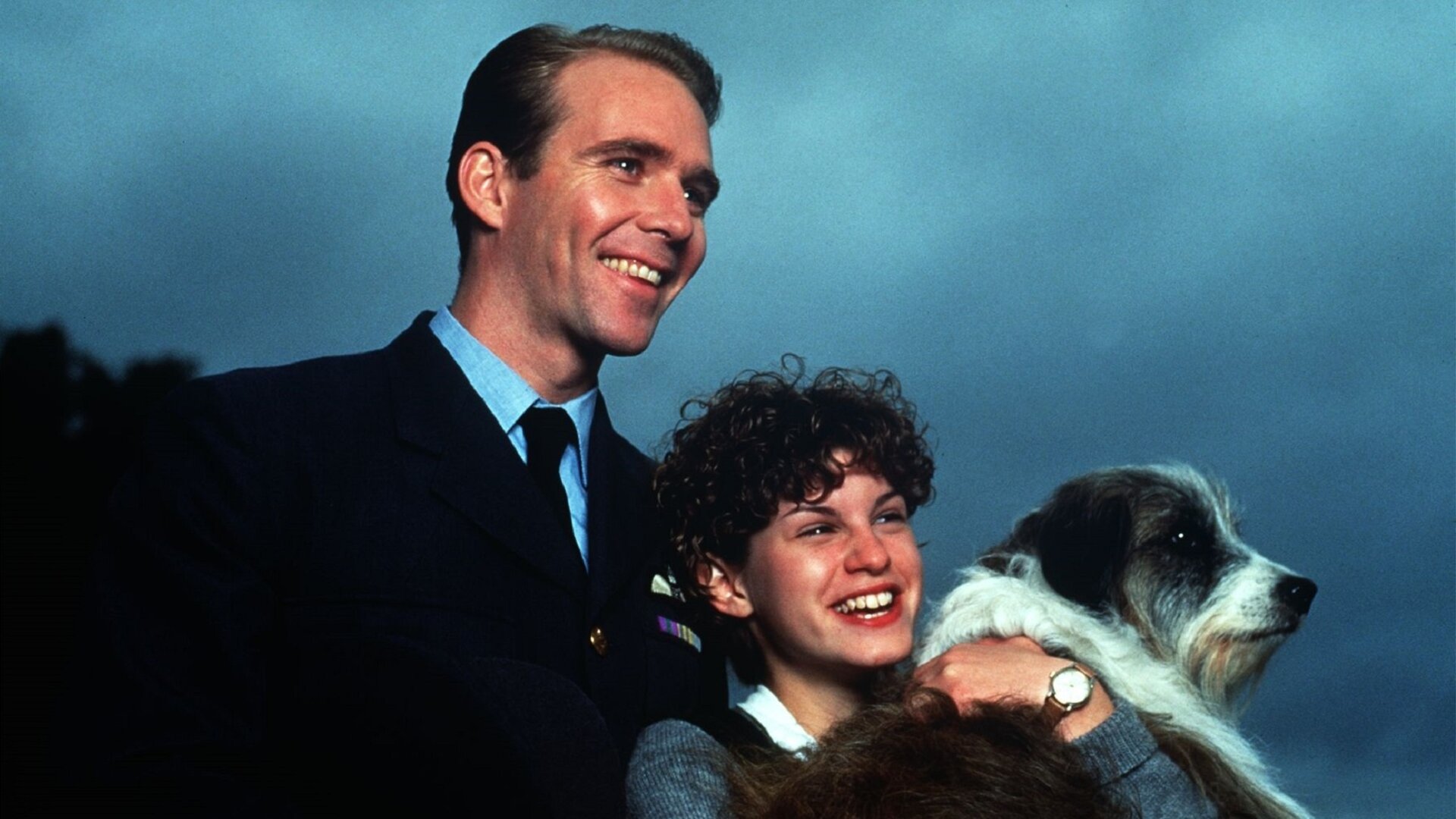 "Fünf Freunde als Retter in der Not": Jeff Thomas (Jason Connery) steht hinter George (Jemima Rooper), die den Hund Timmy im Arm hält. Jeff Thomas (Jason Connery) hat seine rechte Hand auf Georges Arm gelegt. Die beiden lachen und schauen zur Seite.