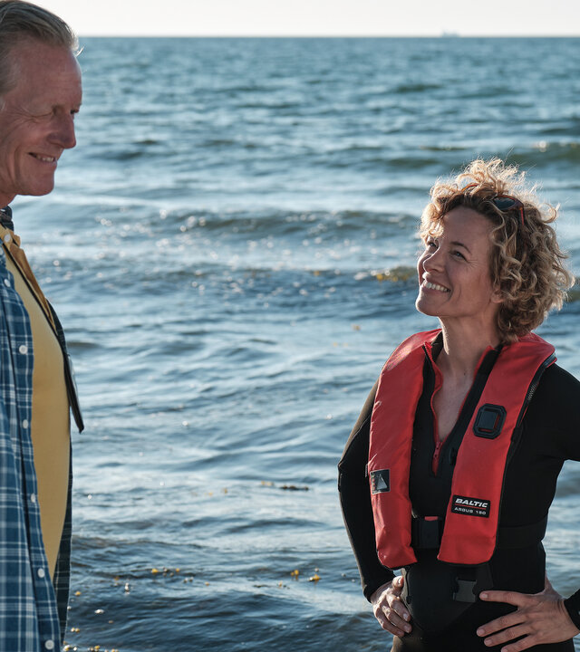 "Dan Sommerdahl - Tödliche Idylle: Das Cannes des Nordens": Dan Sommerdahl (Peter Mygind) und Josefine (Maibritt Saerens) stehen sich gegenüber und lächeln sich an. Im Hintergrund ist das Meer zu sehen.