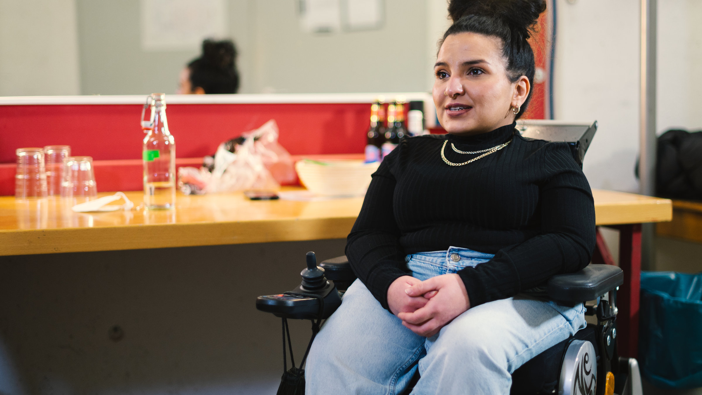 "Einfach Mensch! - 'Wir werden behindert!'": Kübra Sekin, die die Glasknochenkrankheit hat, sitzt in ihrem Rollstuhl vor einem Tisch mit Spiegel und schaut nach links aus dem Bild.