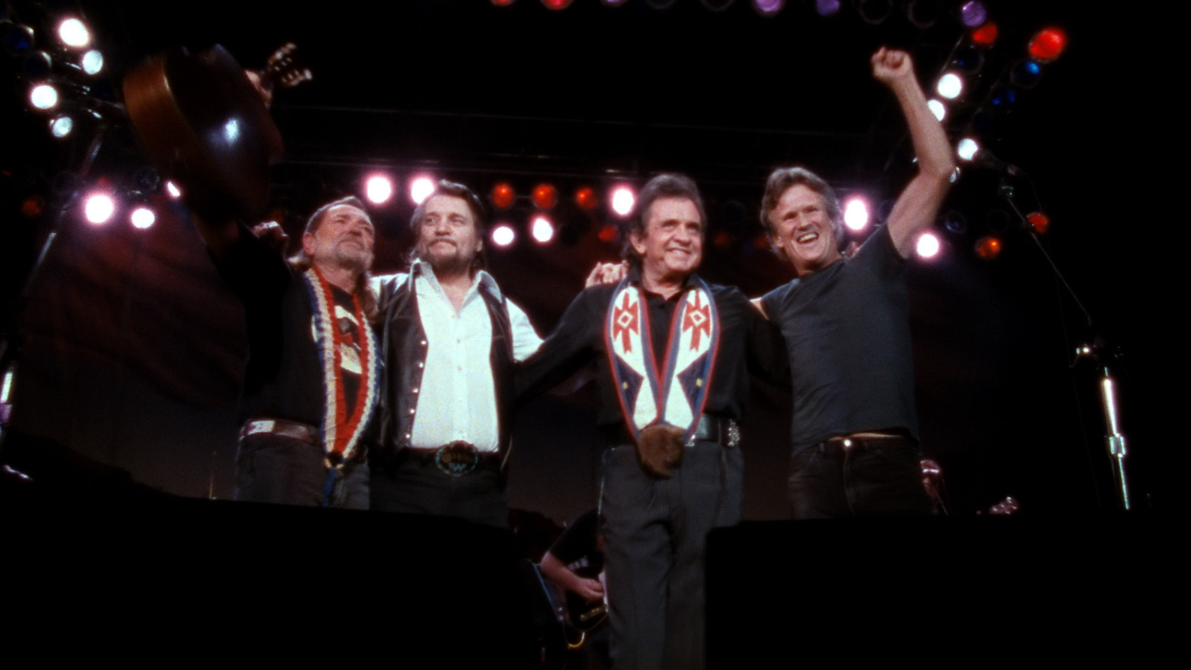 "The Highwaymen: American Outlaws - Live": Vier Bandmitglieder stehen nebeneinander auf einer Bühne, die Arme jeweils um die Schulter des Nebenmannes gelegt. Der Mann rechts außen hat seinen linken Arm nach oben gestreckt.