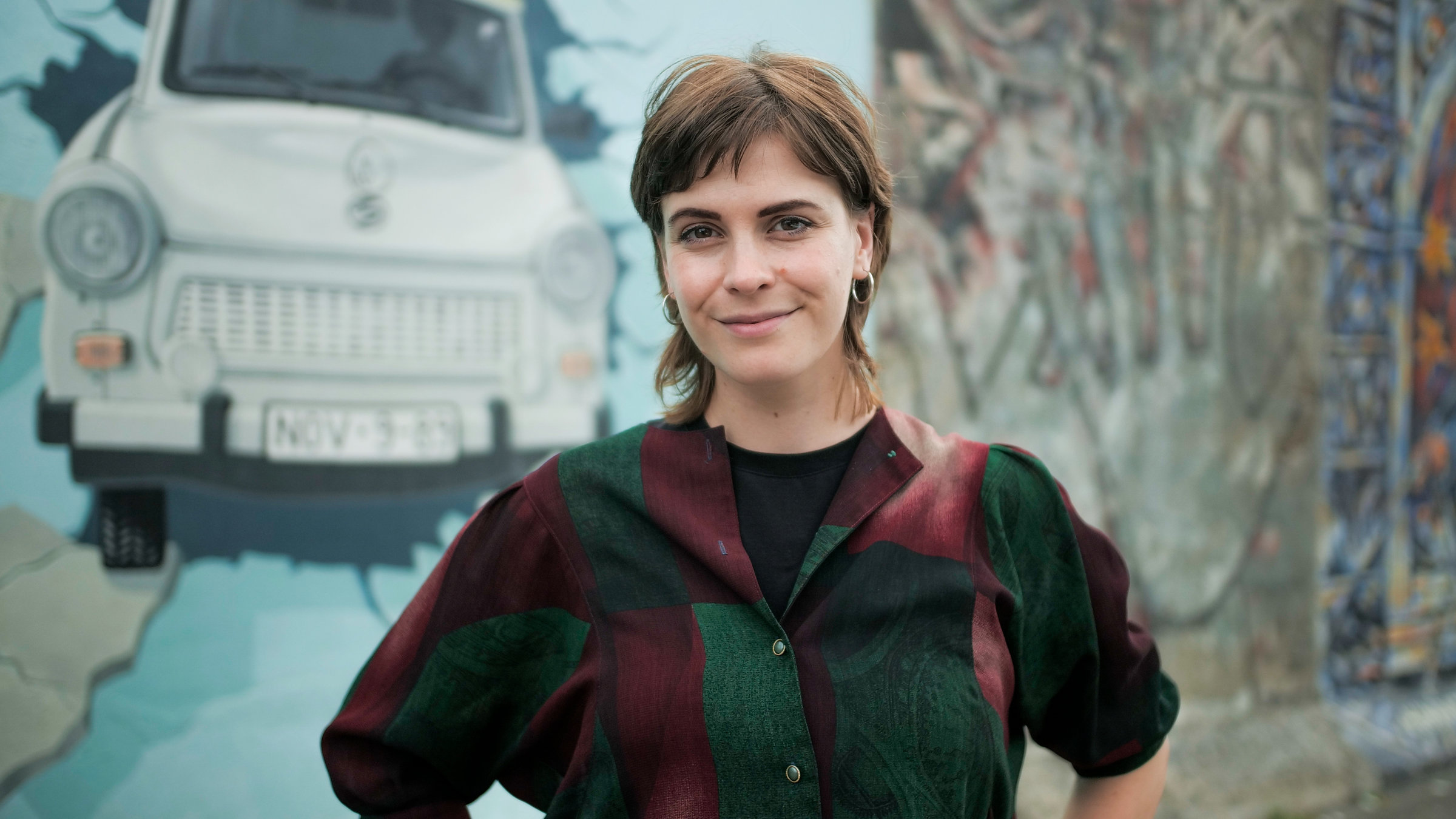 "37° Leben - Ostkinder": Julia Lorenz steht vor einem Stück der Berliner Mauer an der East-Side-Gallery und schaut freundlich in die Kamera. Auf dem Mauerstück sieht man einen Trabi, der die Mauer durchbricht.
