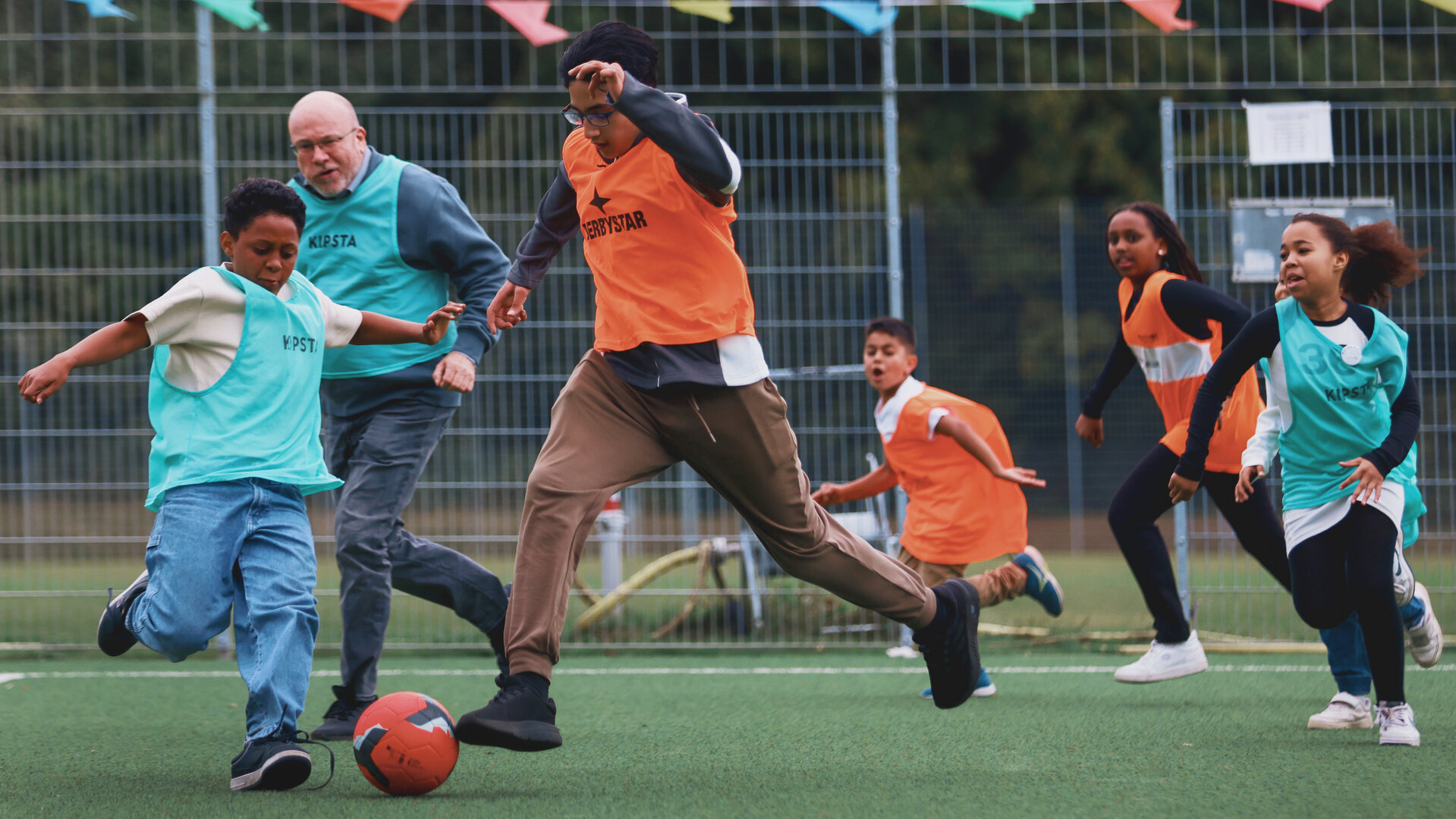 "Aktion Mensch - Glückszahlen der Woche": Auf einem Fußballfeld spielen mehrere Kinder und Erwachsene ein dynamisches Spiel. Die Teams tragen farbige Leibchen in Orange und Türkis.