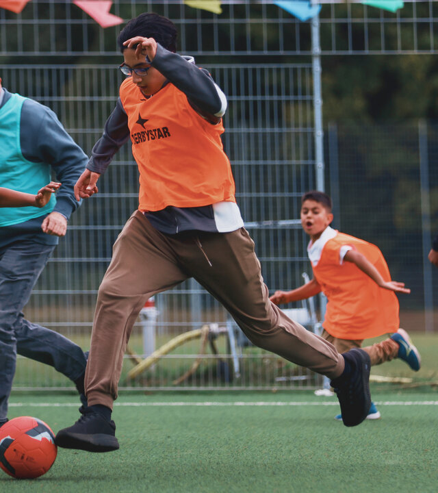 "Aktion Mensch - Glückszahlen der Woche": Auf einem Fußballfeld spielen mehrere Kinder und Erwachsene ein dynamisches Spiel. Die Teams tragen farbige Leibchen in Orange und Türkis.