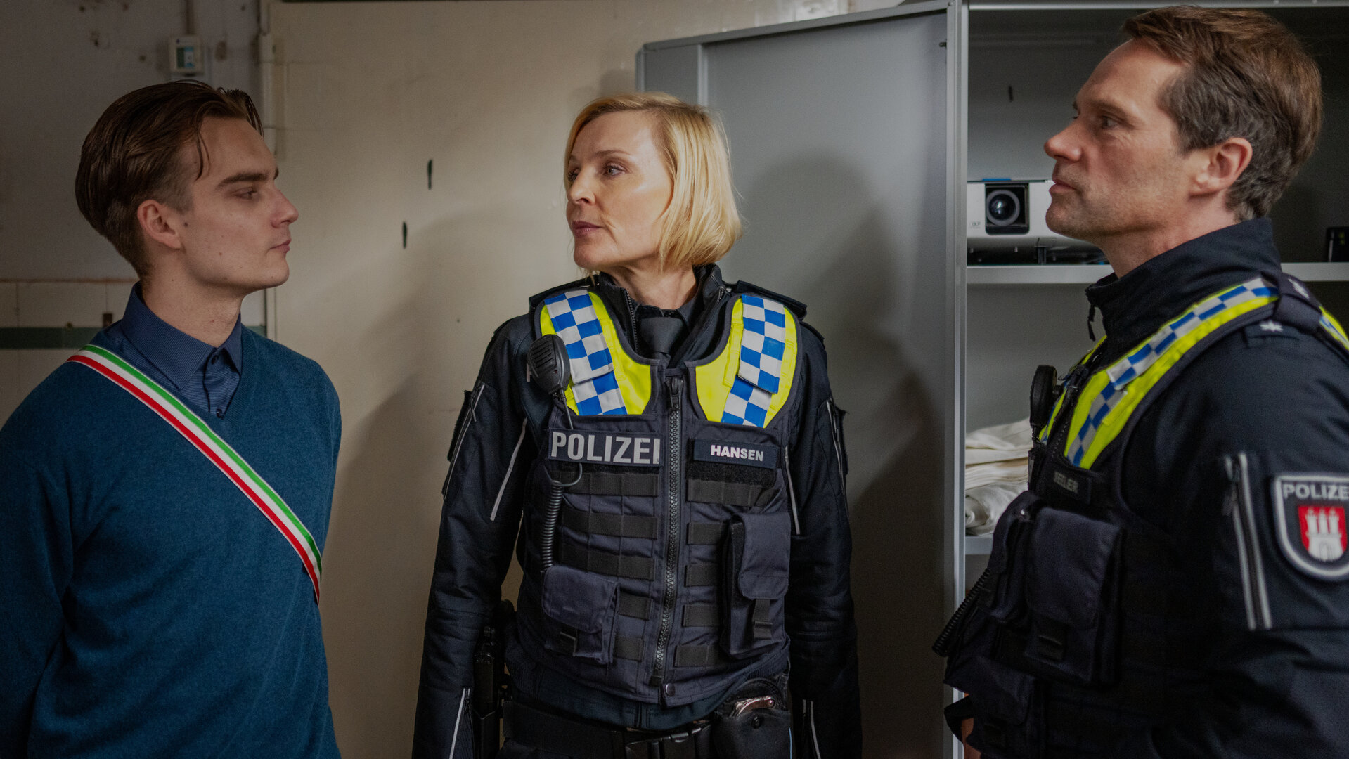 "Notruf Hafenkante - Ehrenwertes Haus": Die Polizistin Melanie Hansen (Sanna Englund) und ihr Kollege Mattes Seeler (Matthias Schloo) stehen mit Marvin (Mateo Wansing Lorrio) in einem Kellerraum. Beide schauen ihn ernst an.
