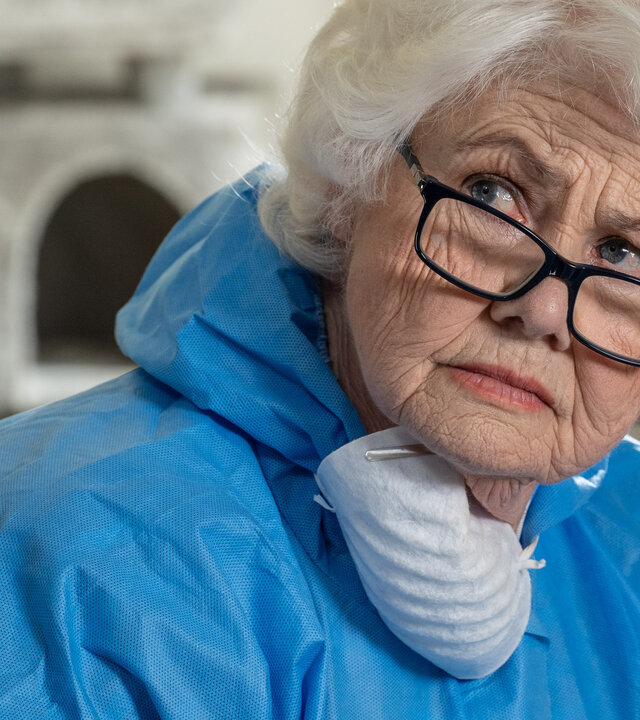 "Inspector Barnaby - Von Hunden und Menschen": Nahaufnahme: Rechtsmedizinerin Fleur Perkins (Annette Badland) trägt einen blauen Ganzkörper-Overall und eine Atemmaske um ihren Hals. Dabei blickt sie ernst nach zur Seite. Im Hintergrund beugt sich ein Forensiker (unbekannter Darsteller) nach unten.