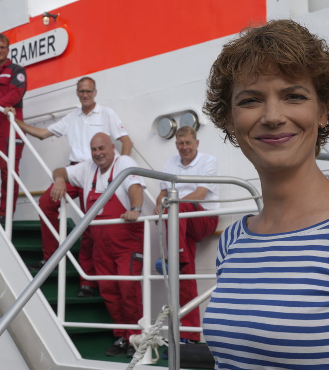 "Terra Xpress: Faszination Nordsee - Mit den Seenotrettern im Einsatz": Moderatorin Lena Ganschow steht, im blau-weiß gestreiften Shirt, vor einem Boot und lächelt in die Kamera. Auf dem Boot sieht man vier Männer in rot-weißer Kleidung, die an der Reling stehen.